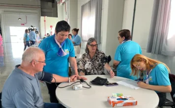 Saúde: Moradores de Arroio do Silva têm manhã de cuidados Profissionais de saúde do Balneário Arroio do Silva, vestindo camisetas azuis, aferindo a pressão arterial de um morador durante o evento "Dia de Cuidar da Saúde" no Centro Multiuso.