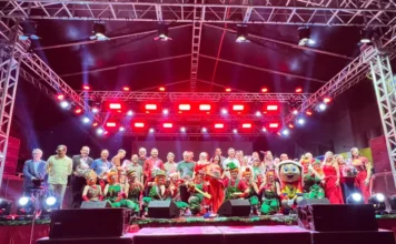 Sombrio abre Natal com chegada do Papai Noel e desfile Grupo de cerca de 40 pessoas no palco principal da festa de Natal em Sombrio. O grupo é composto por autoridades (adultos com trajes sociais), crianças e adultos fantasiados de ajudantes do Papai Noel (elfos em verde e vermelho) e um mascote, sob uma estrutura metálica iluminada com fortes luzes vermelhas.