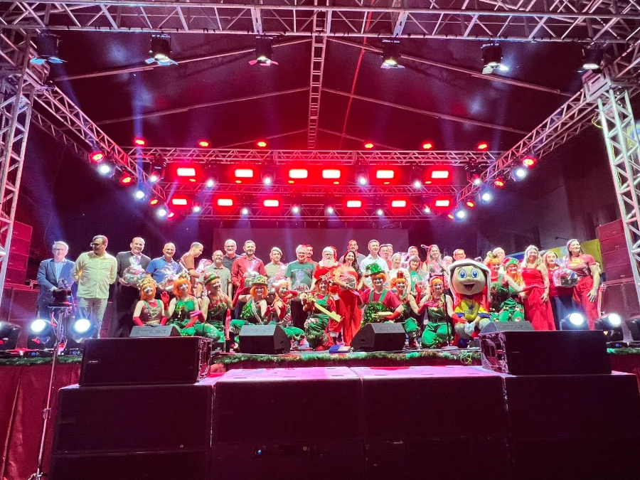 Sombrio abre Natal com chegada do Papai Noel e desfile Grupo de cerca de 40 pessoas no palco principal da festa de Natal em Sombrio. O grupo é composto por autoridades (adultos com trajes sociais), crianças e adultos fantasiados de ajudantes do Papai Noel (elfos em verde e vermelho) e um mascote, sob uma estrutura metálica iluminada com fortes luzes vermelhas.