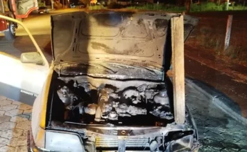 Susto em Turvo: motor de carro pega fogo na madrugada Frente de um Chevrolet Kadett com o capô aberto sustentado por uma madeira, revelando o motor totalmente carbonizado pelo fogo. O veículo está parado sobre paralelepípedos à noite, com parte de um caminhão do Corpo de Bombeiros visível ao fundo.