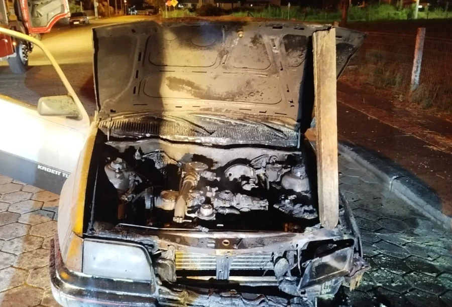 Frente de um Chevrolet Kadett com o capô aberto sustentado por uma madeira, revelando o motor totalmente carbonizado pelo fogo. O veículo está parado sobre paralelepípedos à noite, com parte de um caminhão do Corpo de Bombeiros visível ao fundo.