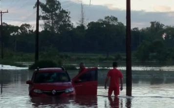 Turvo: Bombeiros resgatam idoso ilhado após rio transbordar Bombeiro Militar em uniforme vermelho em meio à enchente, auxiliando a retirar um idoso de dentro de um carro submerso em água na Estrada Geral de Itoupava II, em Turvo, após o rio transbordar.