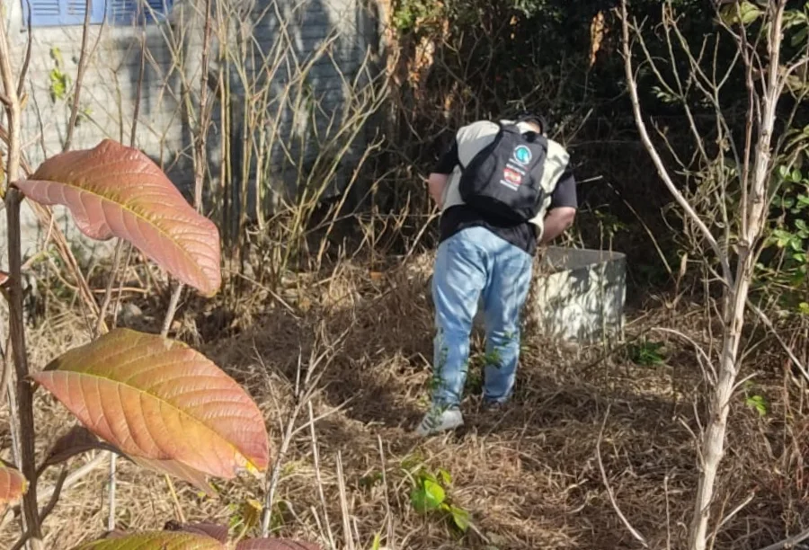 Técnico de endemias de Turvo inspecionando uma área externa com vegetação e um recipiente abandonado, como parte da ação de combate à dengue nas repartições públicas do município.