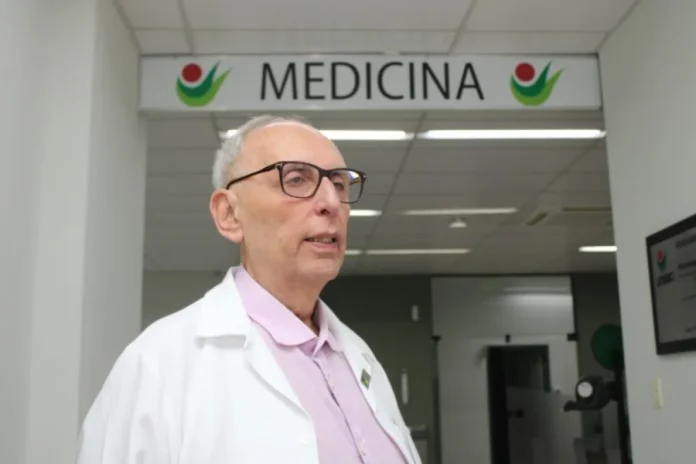 Unesc decreta luto oficial por morte de professor de Medicina Professor de Medicina Dario Henrique Garcia, com jaleco e óculos, em frente à placa do curso na UNESC, cuja morte motivou o decreto de cinco dias de luto oficial na universidade.