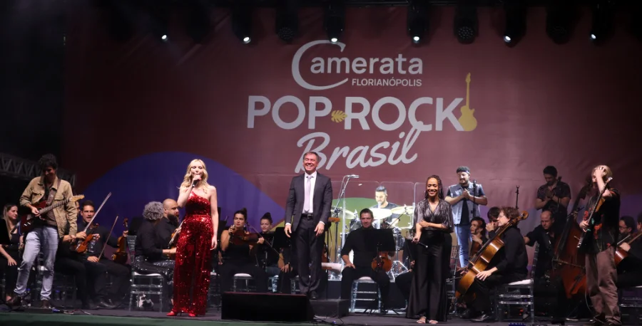 A Camerata Florianópolis se apresenta no palco com orquestra e cantores, sob o banner 