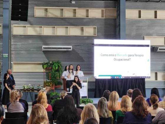 Apresentação em palco de evento universitário, com palestrantes em frente a uma tela que exibe o lançamento dos novos cursos de Fonoaudiologia e Terapia Ocupacional.
