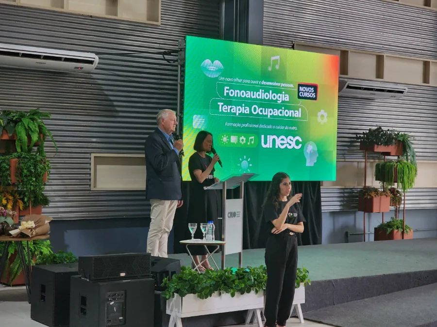 Dois palestrantes (um homem e uma mulher) no púlpito, com uma intérprete de Libras à frente, durante o lançamento dos novos cursos da Unesc.