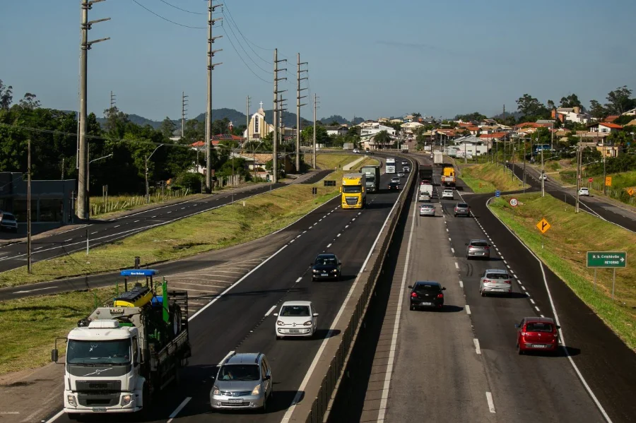 Visão da BR-101 Sul/SC, em pista dupla e movimento, com caminhões e carros circulando, e área urbana ao fundo.