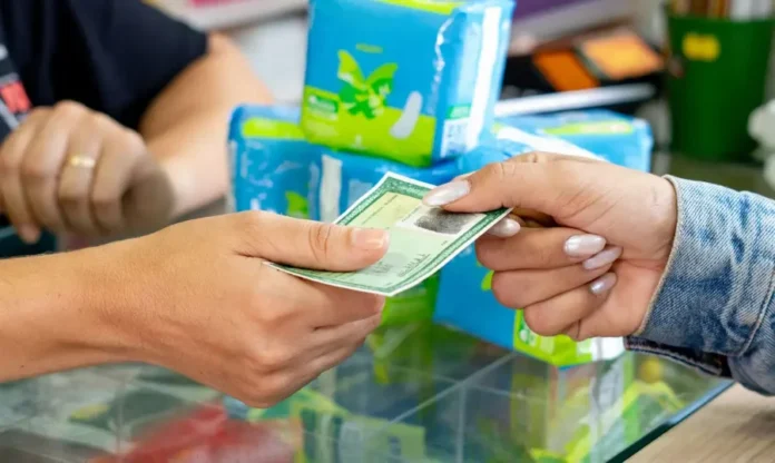 14 milhões de mulheres recebem aviso para retirar absorventes Mãos trocando um documento (Autorização de Retirada) em uma farmácia popular, com pacotes de absorventes azuis em segundo plano.