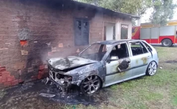 Ação rápida evita que fogo em carro atinja casa em Sombrio Carro (Volkswagen Gol) cinza totalmente destruído por incêndio, encostado em uma parede de tijolos de uma residência, com um caminhão de bombeiros ao fundo.