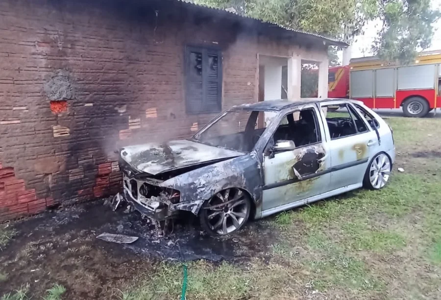Carro (Volkswagen Gol) cinza totalmente destruído por incêndio, encostado em uma parede de tijolos de uma residência, com um caminhão de bombeiros ao fundo.