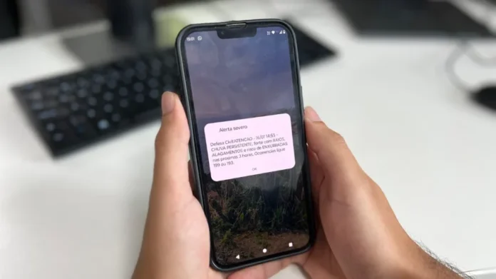 Alerta de tempestade chega a celulares sem cadastro em Sombrio Pessoa segurando um celular que exibe um Alerta Severo da Defesa Civil sobre chuva persistente, forte com raios e risco de alagamentos.