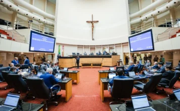 Alesc aprova lei que isenta catarinense da taxa anual do CRLV Imagem do Plenário da Assembleia Legislativa de Santa Catarina (Alesc), mostrando deputados sentados em semicírculo em frente às suas mesas de trabalho com laptops abertos, durante uma sessão de votação. Duas grandes telas exibem informações da sessão no fundo. Uma cruz com a imagem de Jesus Cristo está pendurada acima da mesa diretora, ao centro.