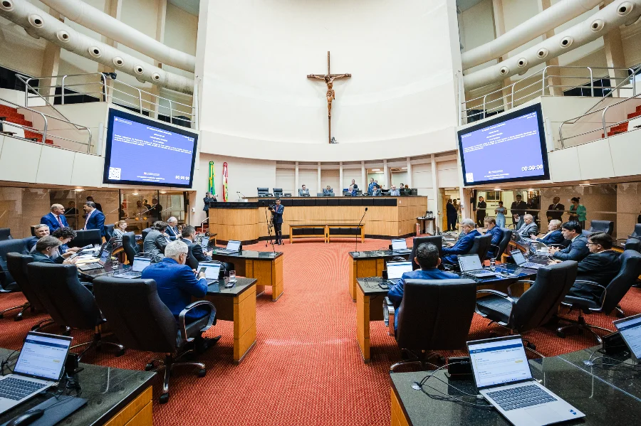 Alesc aprova lei que isenta catarinense da taxa anual do CRLV Imagem do Plenário da Assembleia Legislativa de Santa Catarina (Alesc), mostrando deputados sentados em semicírculo em frente às suas mesas de trabalho com laptops abertos, durante uma sessão de votação. Duas grandes telas exibem informações da sessão no fundo. Uma cruz com a imagem de Jesus Cristo está pendurada acima da mesa diretora, ao centro.