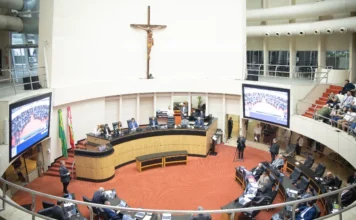Alesc corrige desigualdade de valores para urgência e emergência Foto panorâmica do plenário da Assembleia Legislativa de Santa Catarina (Alesc) visto de cima. Ao centro, destaca-se a mesa diretora em madeira com um grande crucifixo fixado na parede branca ao fundo. Abaixo, os deputados estaduais estão sentados em suas bancadas em formato de semicírculo, utilizando notebooks. Nas laterais, grandes telões exibem a transmissão da sessão.