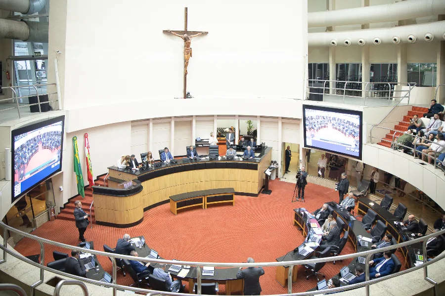 Alesc corrige desigualdade de valores para urgência e emergência Foto panorâmica do plenário da Assembleia Legislativa de Santa Catarina (Alesc) visto de cima. Ao centro, destaca-se a mesa diretora em madeira com um grande crucifixo fixado na parede branca ao fundo. Abaixo, os deputados estaduais estão sentados em suas bancadas em formato de semicírculo, utilizando notebooks. Nas laterais, grandes telões exibem a transmissão da sessão.