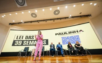 Alesc debate PL que dá direito à cesariana eletiva Deputada Paulinha discursa em um auditório em frente a um grande painel que exibe a mensagem "LEI DAS 39 SEMANAS", com QR code e autoridades sentadas no palco.