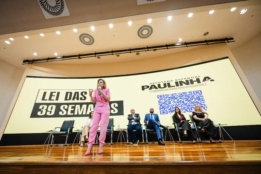 Deputada Paulinha discursa em um auditório em frente a um grande painel que exibe a mensagem 