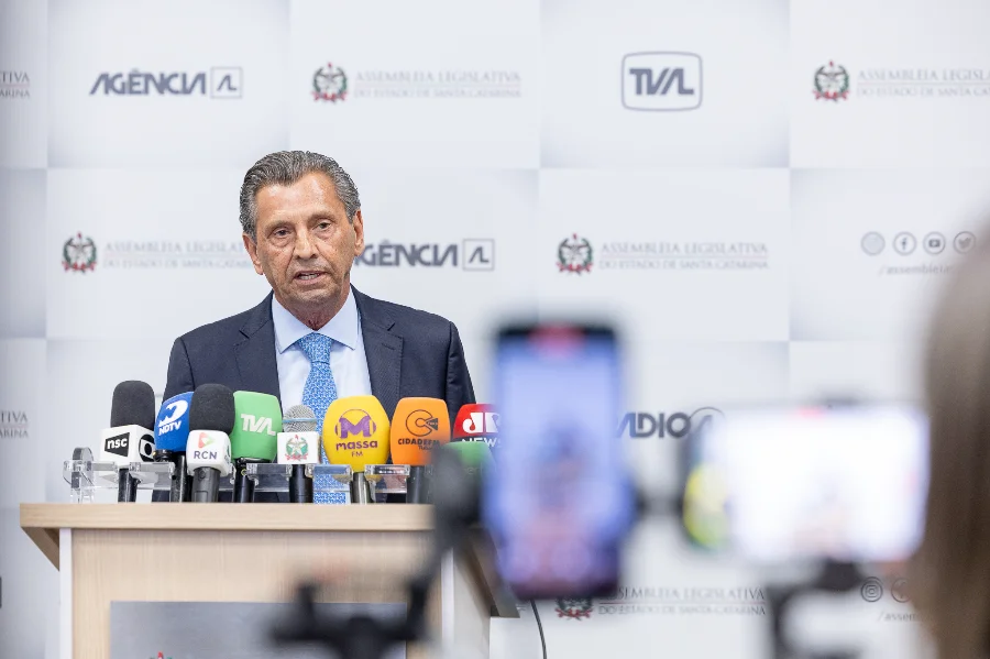 Alesc economiza e devolve R$ 403 milhões ao Governo de SC O presidente da Assembleia Legislativa de Santa Catarina, deputado Julio Garcia, fala em um púlpito cercado por microfones de diversas emissoras de rádio e TV. Ao fundo, um painel branco exibe as logomarcas da Alesc, Agência AL e TVAL. Em primeiro plano, o desfoque de um celular registrando a coletiva de imprensa.