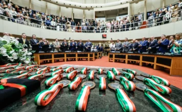 Alesc outorga a 16ª Comenda do Legislativo a 41 homenageados Medalhas da Comenda do Legislativo com fitas nas cores de Santa Catarina (verde, vermelho e branco) dispostas sobre uma mesa em primeiro plano, com o plenário da Alesc lotado e deputados aplaudindo ao fundo.