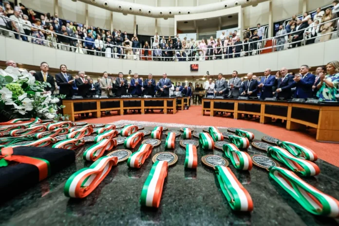 Alesc outorga a 16ª Comenda do Legislativo a 41 homenageados Medalhas da Comenda do Legislativo com fitas nas cores de Santa Catarina (verde, vermelho e branco) dispostas sobre uma mesa em primeiro plano, com o plenário da Alesc lotado e deputados aplaudindo ao fundo.