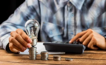 Ar-condicionado e geladeira: Veja como economizar no verão Imagem simbólica de uma lâmpada sobre pilhas de moedas ao lado de uma calculadora, representando dicas da Celesc para economizar na conta de luz durante o verão.