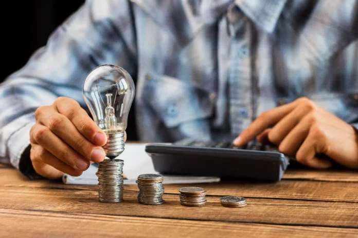 Ar-condicionado e geladeira Veja como economizar no verão Imagem simbólica de uma lâmpada sobre pilhas de moedas ao lado de uma calculadora, representando dicas da Celesc para economizar na conta de luz durante o verão.