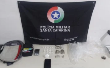 Araranguá: Jovem de 20 anos tenta fugir, mas é preso por tráfico Materiais apreendidos pela Polícia Militar de Santa Catarina (PMSC) expostos sobre uma mesa branca, com uma bandeira preta da PMSC ao fundo. Os itens incluem uma mochila, uma balança de precisão, dois celulares (um smartphone preto e um telefone simples), diversas porções de cocaína e maconha embaladas em sacos plásticos, e materiais para embalagem.