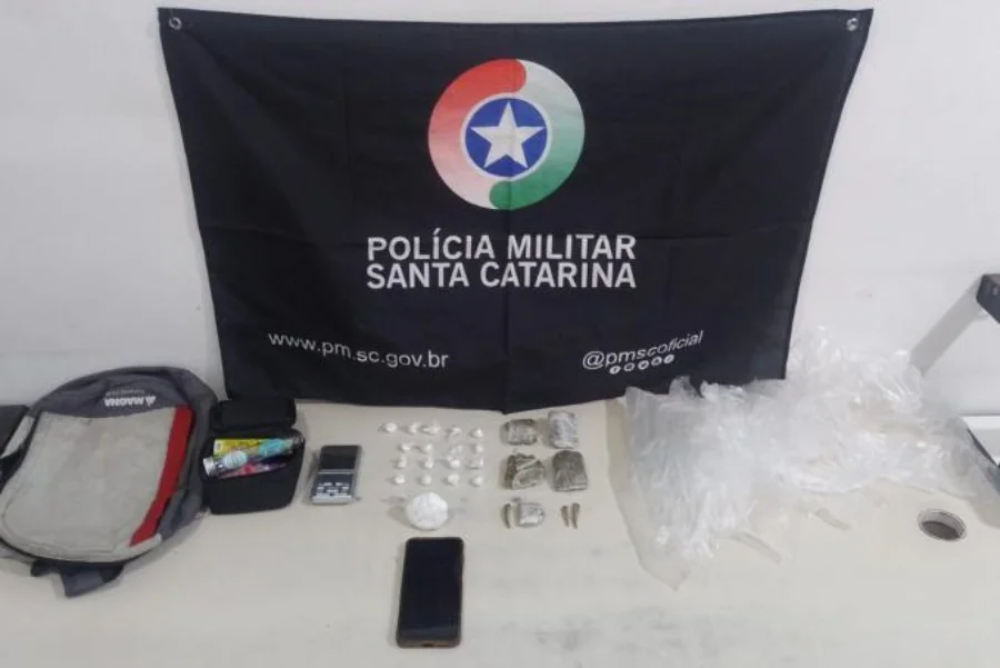 Materiais apreendidos pela Polícia Militar de Santa Catarina (PMSC) expostos sobre uma mesa branca, com uma bandeira preta da PMSC ao fundo. Os itens incluem uma mochila, uma balança de precisão, dois celulares (um smartphone preto e um telefone simples), diversas porções de cocaína e maconha embaladas em sacos plásticos, e materiais para embalagem.