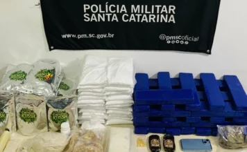Araranguá: PM apreende mais de 65 kg de drogas e insumos Mesa de apreensão com grande quantidade de drogas (maconha, crack) e insumos químicos, dinheiro, celulares e balanças, em frente ao painel da Polícia Militar de Santa Catarina.