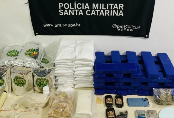 Araranguá PM apreende mais de 65 kg de drogas e insumos Mesa de apreensão com grande quantidade de drogas (maconha, crack) e insumos químicos, dinheiro, celulares e balanças, em frente ao painel da Polícia Militar de Santa Catarina.