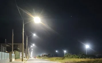 Araranguá instala iluminação LED no Morro dos Conventos Fotografia noturna de uma rua pavimentada na orla do Morro dos Conventos, em Araranguá. Postes de madeira sustentam novas luminárias de LED que emitem uma luz branca intensa, iluminando claramente a via e a vegetação rasteira ao lado. Ao fundo, as luzes se estendem em linha reta pela orla, sob um céu escuro.