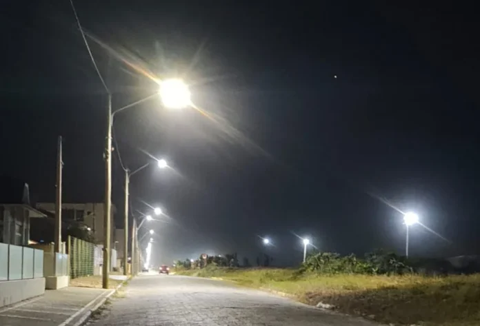 Araranguá instala iluminação LED no Morro dos Conventos Fotografia noturna de uma rua pavimentada na orla do Morro dos Conventos, em Araranguá. Postes de madeira sustentam novas luminárias de LED que emitem uma luz branca intensa, iluminando claramente a via e a vegetação rasteira ao lado. Ao fundo, as luzes se estendem em linha reta pela orla, sob um céu escuro.