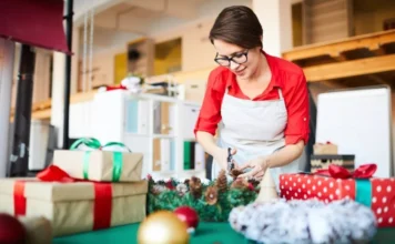 Araranguá terá comércio aberto até 22h a partir de 15 de dezembro Mulher (atendente ou artesã) sorrindo, vestida com camisa vermelha e avental branco, cortando um arranjo de pinhas sobre uma mesa com presentes de Natal embrulhados.