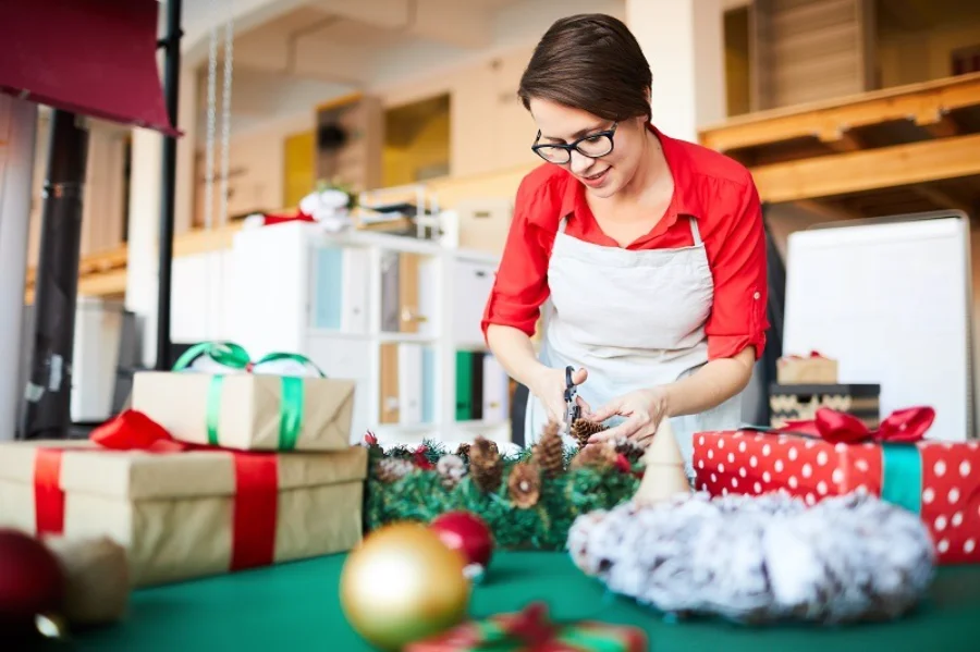 Araranguá terá comércio aberto até 22h a partir de 15 de dezembro Mulher (atendente ou artesã) sorrindo, vestida com camisa vermelha e avental branco, cortando um arranjo de pinhas sobre uma mesa com presentes de Natal embrulhados.