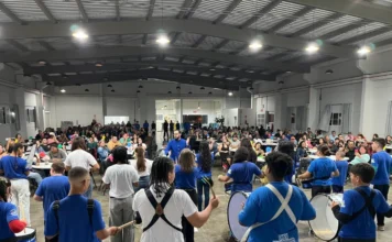 Arroio do Silva: SCFV celebra talentos de 105 jovens e crianças Vista de uma plateia em um grande salão coberto, onde um grupo de crianças e jovens, a maioria vestindo camisetas azuis, se apresenta com instrumentos de percussão, como tambores e caixas. O público está sentado em mesas, observando a apresentação da III Mostra de Talentos do Serviço de Convivência e Fortalecimento de Vínculos (SCFV).