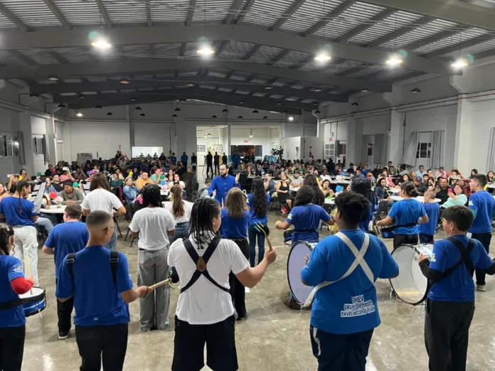 Arroio do Silva SCFV celebra talentos de 105 jovens e crianças Vista de uma plateia em um grande salão coberto, onde um grupo de crianças e jovens, a maioria vestindo camisetas azuis, se apresenta com instrumentos de percussão, como tambores e caixas. O público está sentado em mesas, observando a apresentação da III Mostra de Talentos do Serviço de Convivência e Fortalecimento de Vínculos (SCFV).