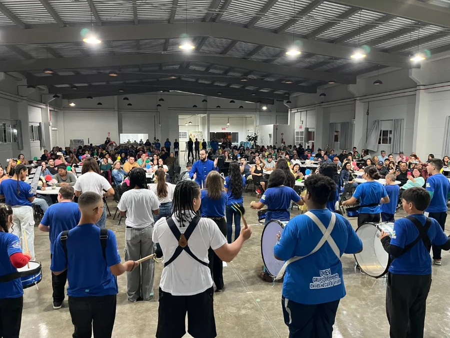 Arroio do Silva SCFV celebra talentos de 105 jovens e crianças Vista de uma plateia em um grande salão coberto, onde um grupo de crianças e jovens, a maioria vestindo camisetas azuis, se apresenta com instrumentos de percussão, como tambores e caixas. O público está sentado em mesas, observando a apresentação da III Mostra de Talentos do Serviço de Convivência e Fortalecimento de Vínculos (SCFV).