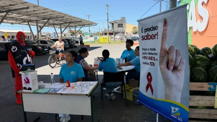 Arroio do Silva intensifica testes rápidos no Dezembro Vermelho Equipe de saúde vestida de azul realizando campanha de testagem rápida de HIV/ISTs em uma tenda em frente a um supermercado em Balneário Arroio do Silva.