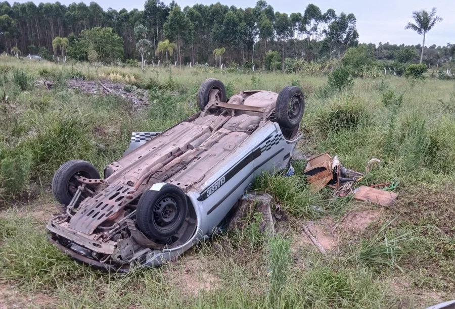 Fotografia de um carro prata modelo sedan capotado de cabeça para baixo em um terreno de vegetação alta e rasteira. Destroços do veículo e objetos pessoais estão espalhados pela grama. Ao fundo, uma linha de árvores de eucalipto sob um céu nublado. O acidente resultou na ejeção de um bebê de um ano.