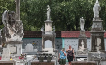 Brasil registra maior aumento de mortes dos últimos 20 anos Vista de um cemitério antigo e arborizado, com grandes lápides e estátuas religiosas em pedestais altos e detalhados. No centro da imagem, duas mulheres (uma em chapéu e blusa azul, a outra de regata preta e short) caminham entre as sepulturas. A foto ilustra o local de registro de óbitos, em referência ao aumento do número de mortes no Brasil.