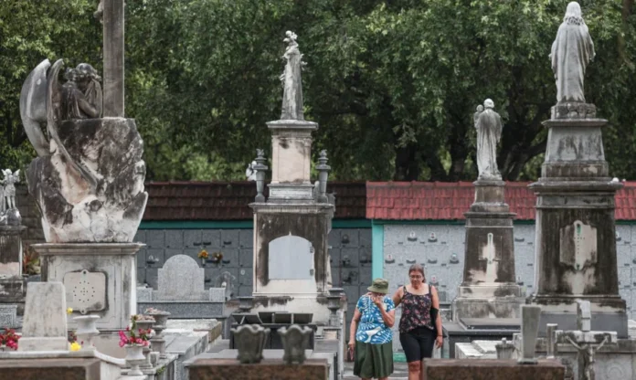 Brasil registra maior aumento de mortes dos últimos 20 anos Vista de um cemitério antigo e arborizado, com grandes lápides e estátuas religiosas em pedestais altos e detalhados. No centro da imagem, duas mulheres (uma em chapéu e blusa azul, a outra de regata preta e short) caminham entre as sepulturas. A foto ilustra o local de registro de óbitos, em referência ao aumento do número de mortes no Brasil.