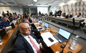 CCJ do Senado aprova projeto que reduz penas do 8 de janeiro Vista ampla do plenário da Comissão de Constituição e Justiça (CCJ) do Senado. Diversos senadores e assessores estão sentados ao longo de grandes mesas de madeira com notebooks. Ao fundo, em uma mesa elevada, o relator Esperidião Amin e outros membros da mesa conduzem a sessão solene. A parede ao fundo possui um painel geométrico em tons de cinza.