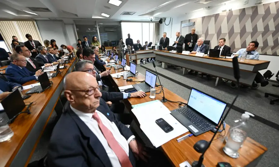 CCJ do Senado aprova projeto que reduz penas do 8 de janeiro Vista ampla do plenário da Comissão de Constituição e Justiça (CCJ) do Senado. Diversos senadores e assessores estão sentados ao longo de grandes mesas de madeira com notebooks. Ao fundo, em uma mesa elevada, o relator Esperidião Amin e outros membros da mesa conduzem a sessão solene. A parede ao fundo possui um painel geométrico em tons de cinza.