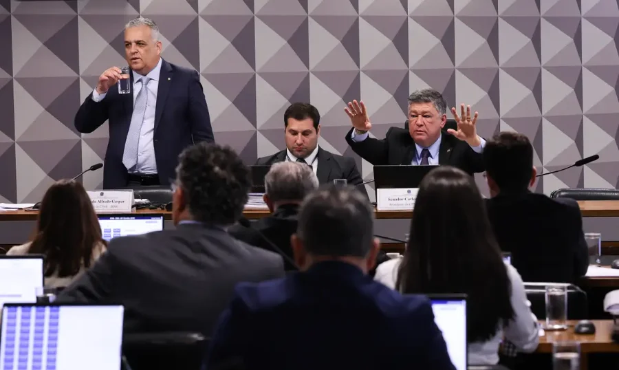 Sessão da CPMI do INSS no Senado, com um político em pé (esquerda) e outro gesticulando (direita), e membros da comissão de costas.