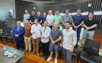 Câmara de Araranguá devolve R$ 4,05 milhões à Prefeitura O presidente da Câmara de Araranguá, vereador Paulinho, entrega um documento oficial ao prefeito César Cesa, simbolizando a devolução de R$ 4,05 milhões. Eles estão cercados por outros vereadores e autoridades no plenário da Câmara, que possui fundo com painéis ripados de madeira.