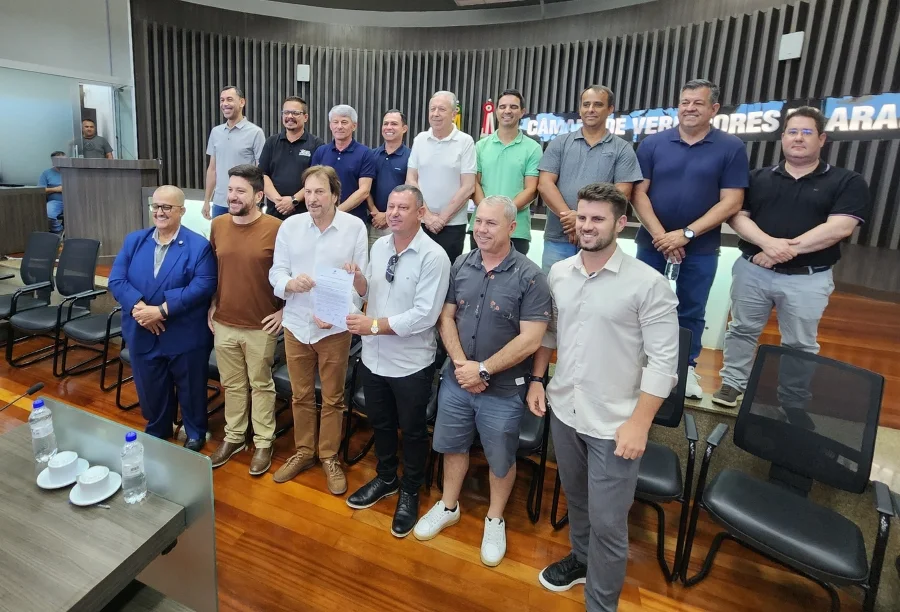 O presidente da Câmara de Araranguá, vereador Paulinho, entrega um documento oficial ao prefeito César Cesa, simbolizando a devolução de R$ 4,05 milhões. Eles estão cercados por outros vereadores e autoridades no plenário da Câmara, que possui fundo com painéis ripados de madeira.