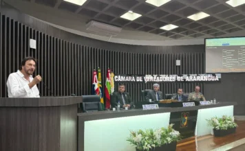 Câmara de Araranguá encerra sessões e Prefeito celebra união Prefeito César Cesa, de camisa social branca e terno preto, em pé no púlpito da Câmara de Vereadores de Araranguá, utilizando um microfone para discursar. Ele gesticula enquanto fala. Ao fundo, três vereadores assistem sentados à mesa diretora, com as bandeiras do Brasil, Santa Catarina e Araranguá ao lado.