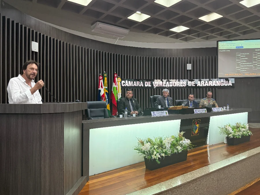 Câmara de Araranguá encerra sessões e Prefeito celebra união Prefeito César Cesa, de camisa social branca e terno preto, em pé no púlpito da Câmara de Vereadores de Araranguá, utilizando um microfone para discursar. Ele gesticula enquanto fala. Ao fundo, três vereadores assistem sentados à mesa diretora, com as bandeiras do Brasil, Santa Catarina e Araranguá ao lado.