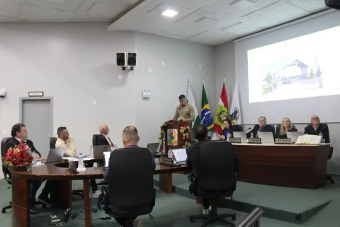 Câmara de Forquilhinha aprova apoio social e ouve balanço da PM Sessão Ordinária na Câmara de Vereadores de Forquilhinha. O Capitão Ramon Marcelina Spillere, de uniforme militar, está em pé em um púlpito de madeira com arranjos de Natal, apresentando o balanço da segurança pública. Os vereadores estão sentados ao redor de uma mesa semicircular em primeiro plano, alguns com notebooks. Ao fundo, uma tela de projeção exibe uma imagem de pórtico da cidade e há quatro bandeiras.